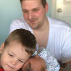 Maintained behaviors image. Photo of a dad looking at his older son holding a new sibling.