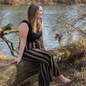 Photo of IABA Owner Jessie Topalov sitting on a log in the woods with a running river in the background.