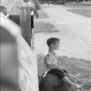 Black and white photo of a young boy sitting a rock outside a farmhouse.